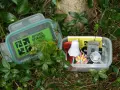 Dwa pojemniki z zawartością do geocaching co to. W jednym zielona naklejka "Official Geocache", w drugim drobne przedmioty jak duch, lupa i "Travel Bug".