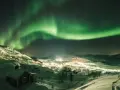 Snöklädda berg och en liten by badar i norrskenets gröna sken. En magisk natt att se norrsken.