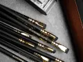 En samling svarta Blackwing-pennor med guldfattningar och suddgummin. En brun läderfodral och en metallbehållare syns också.
