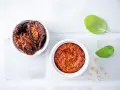 Due ciotoline bianche: una con pomodori secchi interi, l'altra con un invitante pesto di pomodori secchi. Foglie di basilico e pinoli completano la scena.