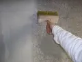 Ręka w białej rękawie nakłada szpachlę na pęknięcia na szarej ścianie za pomocą pędzla.