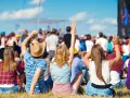 Widok zza pleców na tłum ludzi bawiących się na jednym z letnich festiwali w Polsce. Słońce świeci, niebo jest błękitne.