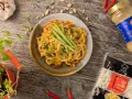 Makaron udon w sosie orzechowym, udekorowany ogórkiem i szczypiorkiem. Obok opakowanie makaronu udon mrożony czy suszony.