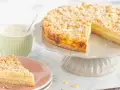 Ein saftiger Streuselkuchen mit Puddingfüllung, bestäubt mit Puderzucker, neben einer Tasse Cappuccino.