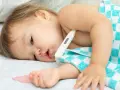 Małe dziecko z termometrem pod pachą, owinięte w kocyk. Wygląda na chore, być może to trzydniówka u dzieci.