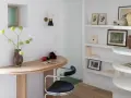 Un despacho pequeño en casa con un escritorio curvo de madera, una silla de terciopelo azul y estanterías blancas llenas de libros y objetos decorativos.