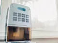 Deumidificatore bianco e azzurro con serbatoio trasparente, posizionato vicino a una finestra. Il deumidificatore fa aria calda, creando un ambiente confortevole.