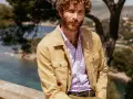 Un uomo con barba e capelli ricci indossa una camicia a quadri viola e una giacca color sabbia. Ottima idea su cosa mettere sopra la camicia uomo per un look casual chic.