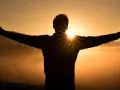 Silhouette eines Mannes mit ausgebreiteten Armen im Sonnenaufgang. Die Sonne bricht durch seine Silhouette und wirft Strahlen. Ein Symbol für Hoffnung und wie wird man Christ.