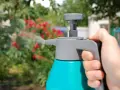 Mano sujetando un pulverizador azul y gris, rociando agua sobre un arbusto con flores rojas. Se puede fumigar en floración para proteger las plantas.