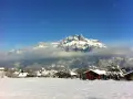 Un pueblo alpino cubierto de nieve, con casas de madera y una iglesia, bajo imponentes montañas nevadas. Un paisaje de invierno en francés.