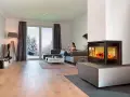 Mujer en sofá mira móvil junto a chimenea de cassette pellets con fuego vivo. Ventana muestra paisaje nevado.
