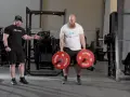 En man med skägg utför farmers walk med tunga vikter. En tränare visar hur övningen ska göras.