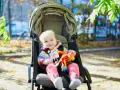 Uśmiechnięte dziecko w kolorowym sweterku i różowych skarpetkach siedzi w spacerówce, trzymając zabawkę. Czas na spacer!
