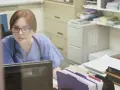 Pielęgniarka w okularach i niebieskim uniformie pracuje przy komputerze, otoczona dokumentami. To miejsce, gdzie zdobywa nowe punkty edukacyjne dla pielęgniarek.