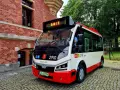 Nowy autobus 100 z napisem "GAIT" na wyświetlaczu, zaparkowany na brukowanej ulicy obok ceglanego budynku.