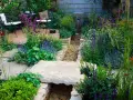 Ein idyllischer **Bachlauf im Garten** mit üppiger Bepflanzung, Steinbrücke und Sitzgelegenheit.