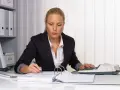 Kobieta w biurze pracuje nad dokumentami, używając kalkulatora i długopisu. Na biurku leżą stosy papierów i segregatorów, a w tle widać regał z dokumentacją. Może przygotowuje weksle.