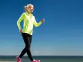 Uśmiechnięta blondynka w neonowej bluzie i czarnych legginsach uprawia power walking nad morzem.