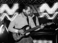 Muzyk gra na gitarze elektrycznej. W tle widać drugiego muzyka i falujące światła, tworzące efekt ekspozycji.
