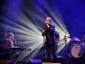 Zespół na scenie, wokalista śpiewa do mikrofonu, klawiszowiec gra na keyboardzie, a perkusista wybija rytm. Wspaniałe tło muzyczne.