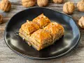 Cztery kawałki baklawy na czarnym talerzu, polane syropem. Obok orzechy włoskie. Idealny baklava przepis na słodki deser.