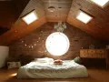 Camera puntata verso un letto in mansarda, con il soffitto in legno e la sua evidente pendenza tetto. Finestre sul tetto e un oblò circolare illuminano la stanza.