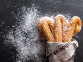 Churros z czekoladą kontra pączki z lukrem? Te złociste churros, posypane cukrem pudrem, kuszą na czarnym tle.