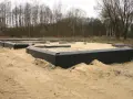Fundament domu w piasku, widoczne czarne membrany izolacyjne i drewniane elementy szalunku. Warstwy gruntu przygotowane do dalszych prac budowlanych.