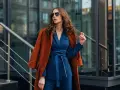 Une femme porte un gilet avec une robe bleu marine, un manteau orange sur les épaules, et des lunettes de soleil.