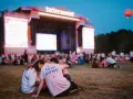 Para siedząca na trawie przed sceną festiwalową z napisem "bittersweet". W tle tłum ludzi i wielkie ekrany.