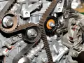 Widok z bliska na mechanizm rozrządu w samochodzie, z widocznymi paskami, kołami zębatymi i napinaczami.