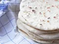 En hög med nybakade tortillabröd, perfekta för ditt nästa tortilla recept. Bredvid ligger en blåvitrutig kökshandduk.