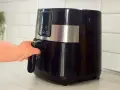 En hand drar ut korgen på en svart airfryer. Den här köksapparaten är lika praktisk som en diskmaskin för snabb matlagning.