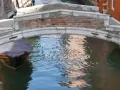 Un piccolo ponte a Venezia, senza parapetto, sovrasta un canale dove una barca coperta attende.