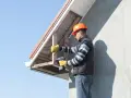 Budowlaniec montuje podbitkę ze styropianu, przykręcając drewniane elementy do okapu domu.