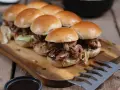 Saftiges pulled chicken im Backofen zubereitet, serviert in kleinen Brötchen auf einem Holzbrett.