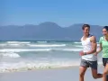 Para biegaczy na plaży, czerpiących energię jak mitochondria co to, z falującym morzem i górami w tle.
