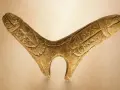 Starożytny, rzeźbiony w drewnie fallus, przypominający stylizowanego ptaka lub zwierzę, z wyrytymi detalami.