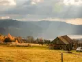 Jesienne słońce przebija się przez chmury nad hrobcza łąka. Stara chata, drzewa w złotych barwach i jezioro w oddali.