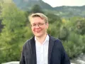 Ein Mann mit Brille lächelt in die Kamera. Er trägt ein weißes Hemd und eine dunkle Jacke. Im Hintergrund sind Bäume und Berge zu sehen. Er studiert Seelsorge.