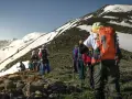 Un groupe de randonneurs progresse sur un sentier rocailleux, le soleil éclairant les montagnes enneigées. Ils semblent déterminés à savoir son altitude.
