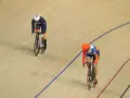 Two cyclists compete in a track sprint. One rider in a blue and white uniform leads, while another in orange and blue follows closely.