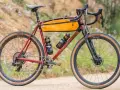 A red gravel bike with an orange frame bag and two water bottles. This bike, like many gravel bikes, does not have suspension, relying on its tires and frame for comfort.
