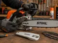 Ręce w rękawiczkach Stihl regulują naciąg łańcucha piły elektrycznej. Obok leży śrubokręt i osłona prowadnicy.