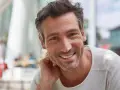 Ein Mann mit einem gepflegten 3-Tage-Bart lächelt in die Kamera. Er sitzt entspannt im Freien.