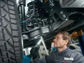 Mechanik bada elementy zawieszenia samochodu, oświetlając je latarką. Widoczna sprężyna i amortyzator.