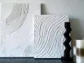 Två vita texturerade tavlor, skapade med spackel, med vågiga mönster. En mörk, taggig skulptur och en rutig ljusbrun och vit ljuslykta står bredvid.