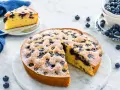 Deliziosa torta allo yogurt con mirtilli, spolverata di zucchero a velo. Una fetta è già servita su un piattino.