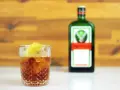 Jägermeister z pomarańczą to świetny drink. Na zdjęciu drink z lodem i plasterkiem pomarańczy obok butelki Jägermeister.